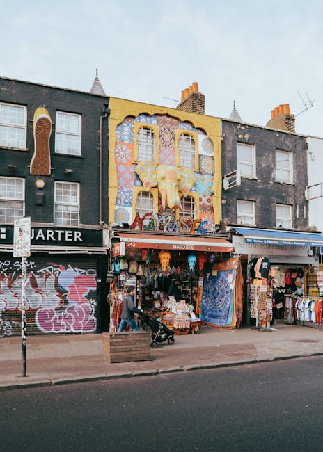 A row of three-story commercial buildings in Kentish Town with graffiti-covered black facades on either side of a central building featuring a large, colorful mural of a smiling elephant with floral patterns, set against a bright yellow background. The ground level showcases various small retail shops with outdoor displays, including clothing racks, bags, and decorative items. In front of the shops, the pavement is occupied by pedestrians, one pushing a baby stroller, and a large wooden planter box. Part of the street is visible with a parked vehicle and street signage, while the upper floors have multiple windows, some open or adorned with window-mounted air conditioning units. The scene depicts a busy urban environment suitable for house removals, with the building facades and sidewalk indicating typical moving logistics preparedness in a city setting.