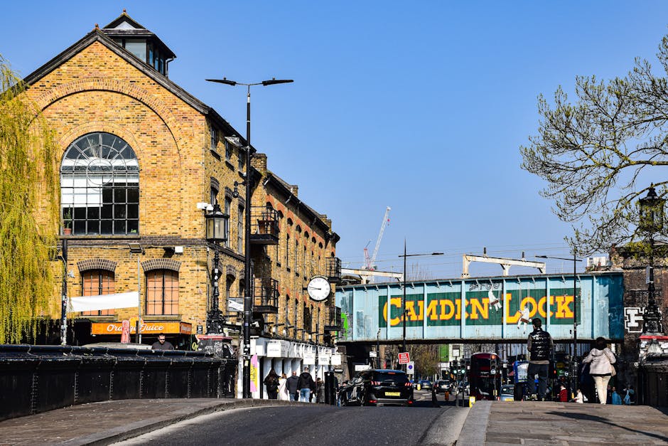 A row of three-story commercial buildings in Kentish Town with graffiti-covered black facades on either side of a central building featuring a large, colorful mural of a smiling elephant with floral patterns, set against a bright yellow background. The ground level showcases various small retail shops with outdoor displays, including clothing racks, bags, and decorative items. In front of the shops, the pavement is occupied by pedestrians, one pushing a baby stroller, and a large wooden planter box. Part of the street is visible with a parked vehicle and street signage, while the upper floors have multiple windows, some open or adorned with window-mounted air conditioning units. The scene depicts a busy urban environment suitable for house removals, with the building facades and sidewalk indicating typical moving logistics preparedness in a city setting.