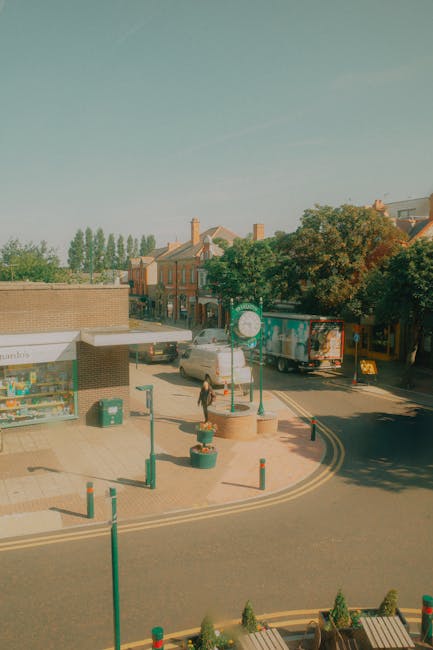 An image capturing a street scene during daytime showing the loading process for a home relocation. In the foreground, there are decorative plant pots and bollards on the pavement, with a woman walking across the street. To the left, a small retail shop with a white awning displays books and magazines, and a green trash bin is placed nearby. In the middle ground, there is a parked silver van and a larger moving truck with company branding, positioned at the curb, likely part of a furniture transport operation handled by Kentish Town Movers. The background features residential buildings with brick facades, a tree with dense foliage providing partial shade, and a clear sky overhead. This setting illustrates the logistical aspect of residential removals, including loading and packing materials, in an urban environment aligned with a professional removals service on Kentish Town Road.
