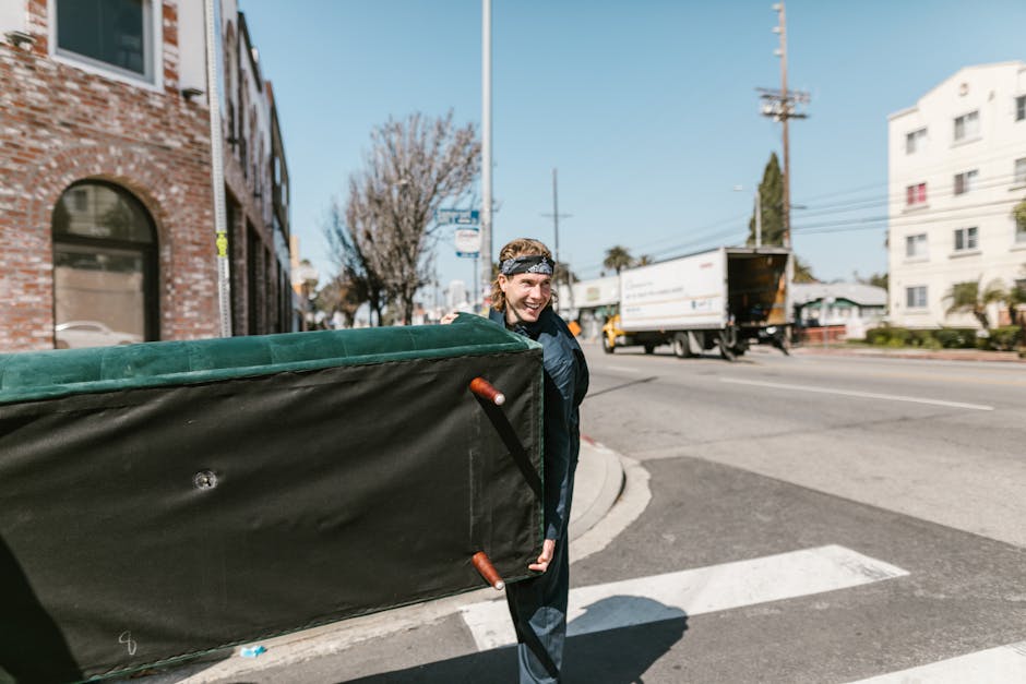 A person with shoulder-length hair, wearing a black jacket and a patterned headband, is outdoors on a city street carrying a large, black padded moving blanket with green edges, used for protecting furniture during a house relocation. The individual is smiling and walking towards a loading area adjacent to a brick building with arched windows on the left. In the background, a white delivery truck with its rear open for loading is visible, along with other vehicles on the road, including a yellow truck and several cars. The pavement has a white crosswalk, and nearby trees are leafless, indicating a possible winter or early spring period. Overhead, there are utility poles with wires and a blue sky with some clouds. The scene is part of a moving process, possibly organized by Kentish Town Movers, featuring elements like packing materials, furniture protection blankets, and loading activities aligned with professional removals and furniture transport services.