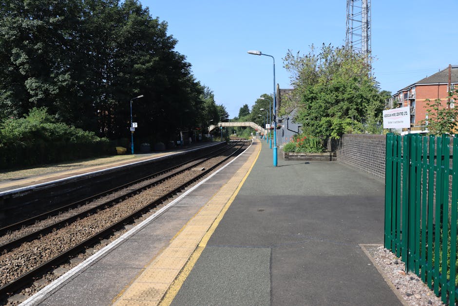 A quiet train station platform with a single set of railway tracks running parallel to the edge of the pavement, which is bordered by a yellow safety line. The platform surface is made of asphalt with tactile paving near the edge. On the right side, there is a green metal fence with vertical bars next to a small garden area with shrubs and trees. A sign on the fence displays the station name, which is not fully visible. Several tall street lamps line the platform, and an overhead bridge or canopy in the distance crosses over the tracks. The scene is well-lit with natural sunlight, and the sky appears clear, indicating a bright day. The station is adjacent to residential buildings with brick facades, and there are no trains or people visible in the image. Kentish Town Movers could facilitate the logistics involved in home relocation through such station environments, supporting efficient furniture transport and packing activities for flats near Kentish Town Station.