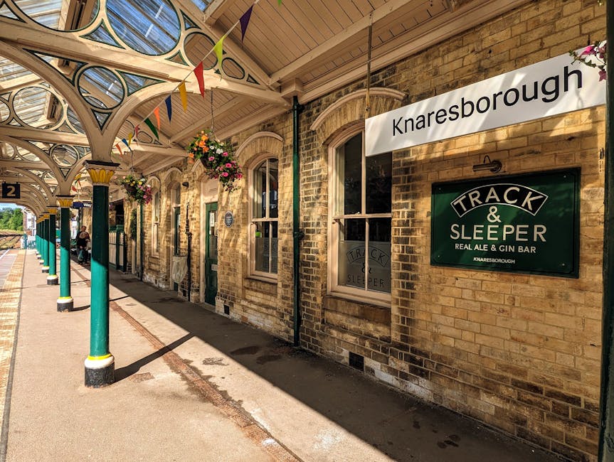 Exterior view of Knaresborough train station platform with a covered waiting area supported by decorative green and cream painted columns, a row of hanging flower baskets, and brick walls featuring arched windows. A sign reading 'Knaresborough' is visible, along with a green sign for 'Track & Sleeper Real Ale & Gin Bar,' indicating the station's amenities. The platform surface is tarmac, with a yellow safety line along the edge, and sunlight casting shadows on the ground. Visible in the background are a few passengers sitting on benches and the station's tracks. The scene captures the station's historical brick architecture and outdoor waiting environment, relevant to home relocation and furniture transport logistics supported by Kentish Town Movers as part of their removals and moving services.