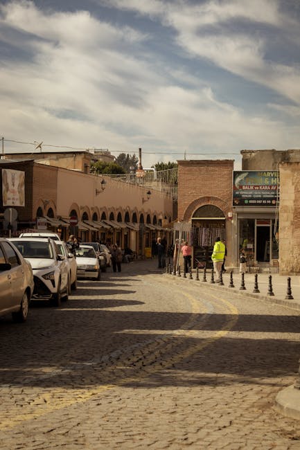 A cobbled street in a busy outdoor marketplace, with a row of retail shops on the left side displaying awnings and signage. Several parked cars are positioned along the street, while pedestrians walk along the pavement, some engaging with shop entrances. An individual wearing a high-visibility vest stands near a gated archway entrance, possibly overseeing movement or providing assistance related to home relocation or logistics. The scene is illuminated by natural daylight, with a partly cloudy sky overhead. The environment suggests an active urban area where furniture or boxes may be loaded or unloaded, reflecting typical aspects of house removals or moving services often coordinated by companies like Kentish Town Movers.