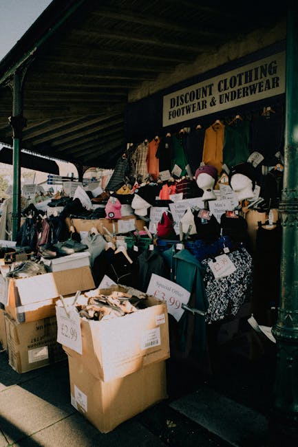 An outdoor market stall under a roofed structure displaying a variety of second-hand clothing, footwear, and accessories for discount sale, with items arranged on tables, in boxes, and hanging from a clothing rack. The footwear includes shoes and boots placed on boxes and the table, while hats and headgear are also visible among the clothing. Several cardboard boxes filled with assorted items are positioned at the front, some with handwritten price tags. Signage above the stall reads 'DISCOUNT CLOTHING FOOTWEAR & UNDERWEAR,' indicating the sale category. The environment appears to be part of a street market, with partial view of the pavement and support pillars. This scene is relevant to relocation services offered by Kentish Town Movers, illustrating the packing or transportation phase of a house move involving clothing and household items, captured in natural daylight.