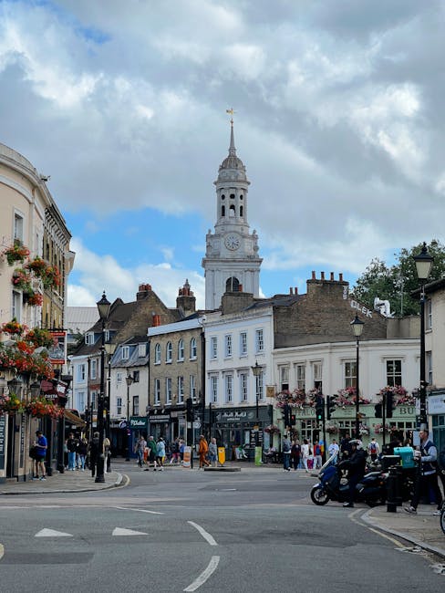 A busy street in Kentish Town with a mix of pedestrians walking along the pavement and vehicles including a scooter and cars moving through the intersection. In the background, there is a prominent historic clock tower with a spire topped by a weather vane, set against a partly cloudy sky. Surrounding the street are buildings with commercial frontages, some adorned with hanging flower baskets and signage. The crowd appears to be engaged in shopping, walking, or casual activities, indicative of day-to-day urban life. The scene reflects typical urban activity associated with house removals and furniture transport, with potential involvement of moving companies like Kentish Town Movers handling equipment and packing materials in this lively neighbourhood setting.