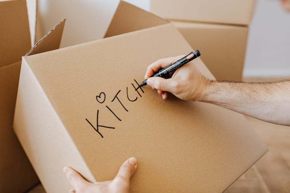 A person’s hand is visible holding a black marker and writing the word 'KITCHEN' on a large cardboard moving box in a home setting. The box is positioned on the floor among other similar cardboard boxes, some of which are partially visible around it. The boxes appear to be part of house packing materials, with the word 'KITCHEN' indicating the contents designated for this particular box. The background shows a neutral-coloured wall and a wooden floor, suggesting an indoor environment prepared for a home relocation process. The image captures the packing stage during furniture transport and moving logistics, with focus on organized packing and labeling for an efficient move, highlighting best practices in packing and loading for a house removal service like those offered by Kentish Town Movers.