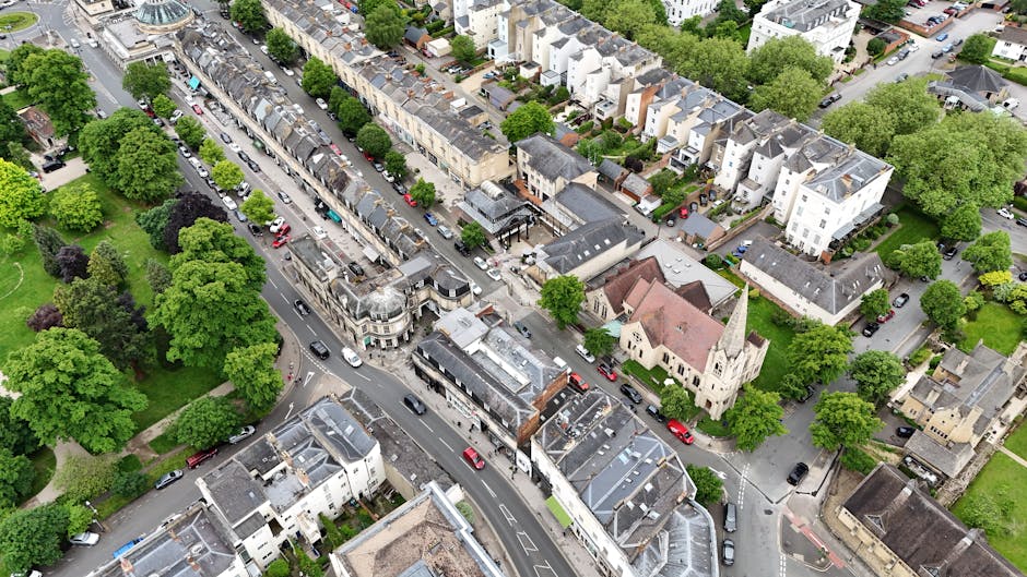 An aerial view of a residential neighbourhood in Kentish Town, featuring a mix of terraced houses, apartment buildings, and a historic church with a tall steeple. The streets are lined with numerous green trees, creating a lush environment. Several cars are parked along the roads and on driveways, with some vehicles moving through the narrow streets. In a nearby driveway, a moving van is parked for a house relocation, with large cardboard boxes, wooden furniture pieces, wrapped and padded items, and packing materials visible as part of the furniture transport process. The scene illustrates a typical urban setting during a home removal, with property access points and the logistics involved in packing and loading household items for transport. The environment is well-lit with natural daylight, capturing the details of the moving process occurring within a tranquil, tree-lined residential area.