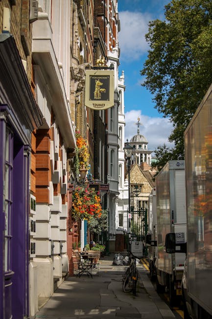 A narrow urban street scene in Kentish Town with parked cars lining the right side of the cobblestone road, including a dark blue hatchback closest to the camera. On the left, there is a sidewalk with